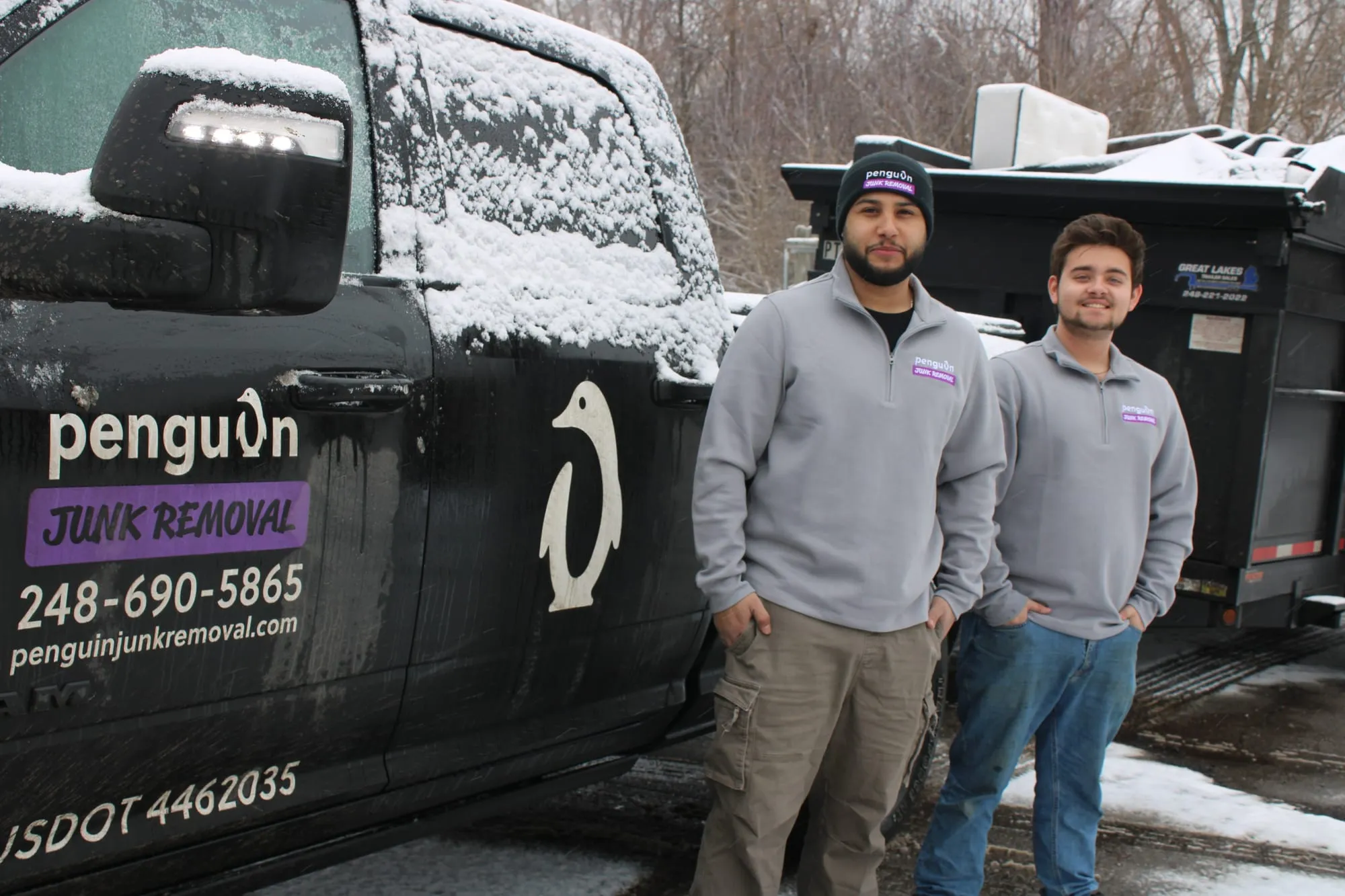 Two uniformed Penguin Junk Removal crew members standing beside their branded truck and dump trailer in Metro Detroit, MI